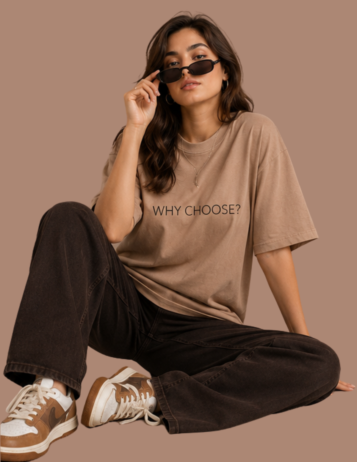 A woman sits cross-legged against a solid taupe background, wearing an oversized beige graphic t-shirt with the text 'WHY CHOOSE?' printed in black, dark brown wide-leg jeans, and brown and white sneakers.