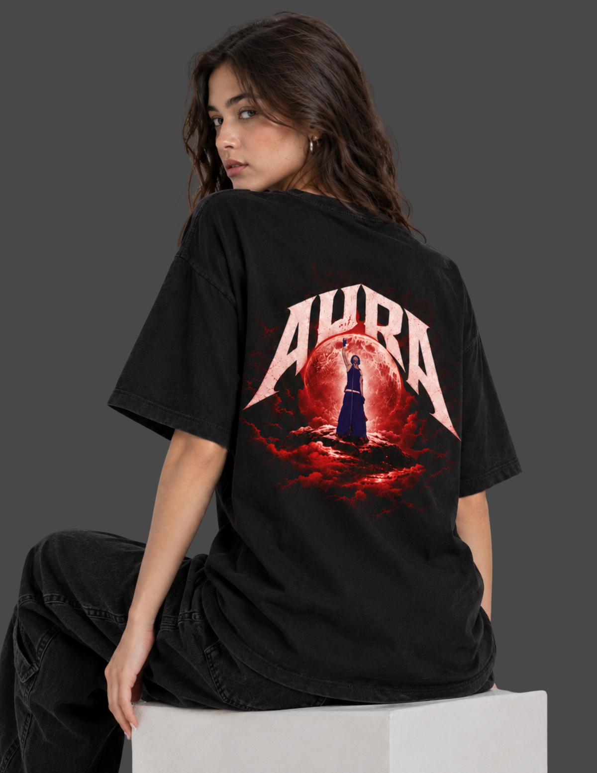 A woman wearing a black oversized T-shirt with a large back graphic featuring the word 'AURA' in white over a red circular design, seated on a white block against a dark gray background.