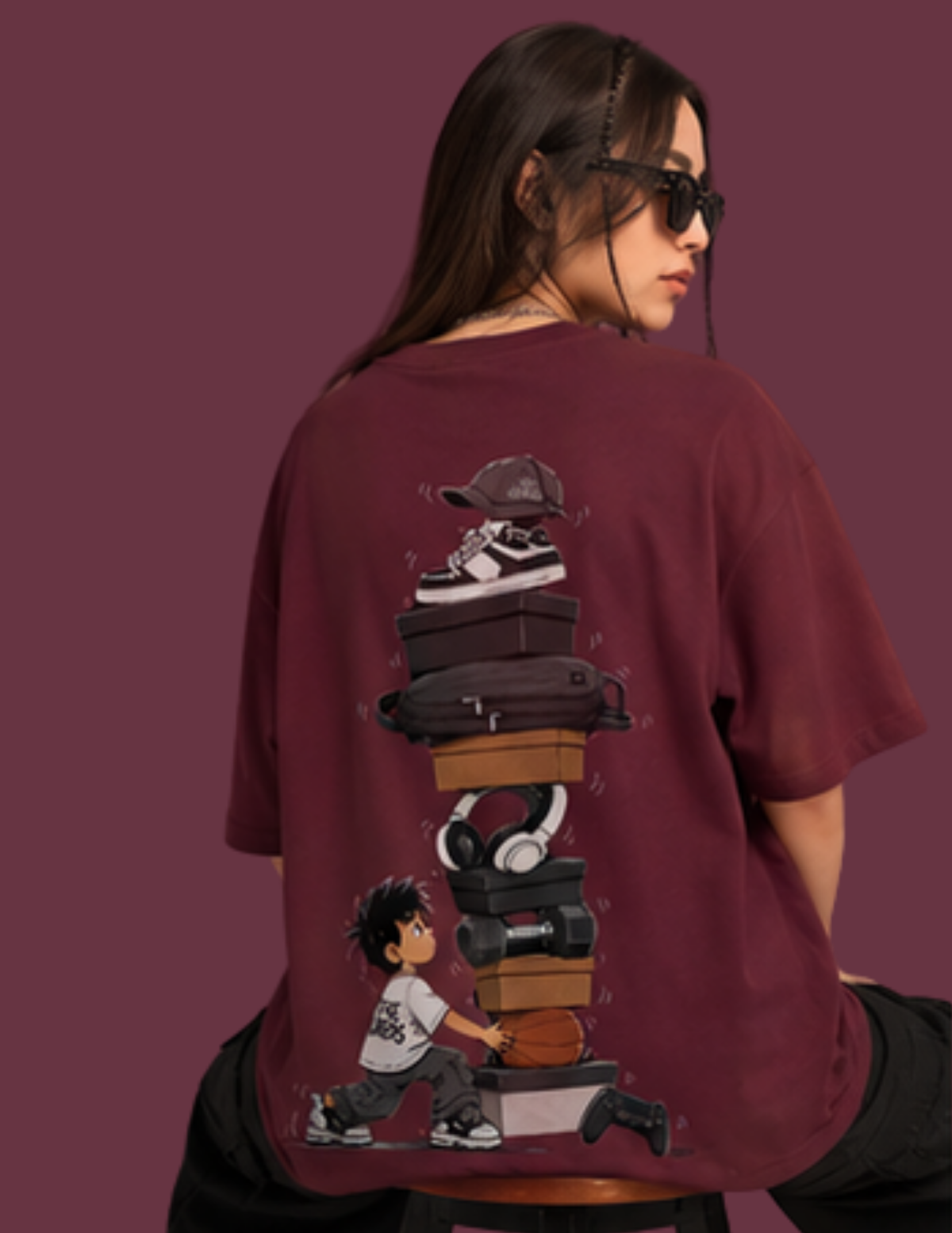 A woman wearing a maroon oversized t-shirt, viewed from the back. The shirt features a large graphic print of a stack of suitcases, headphones, sneakers, and a boy pushing a suitcase, set against a solid maroon background.