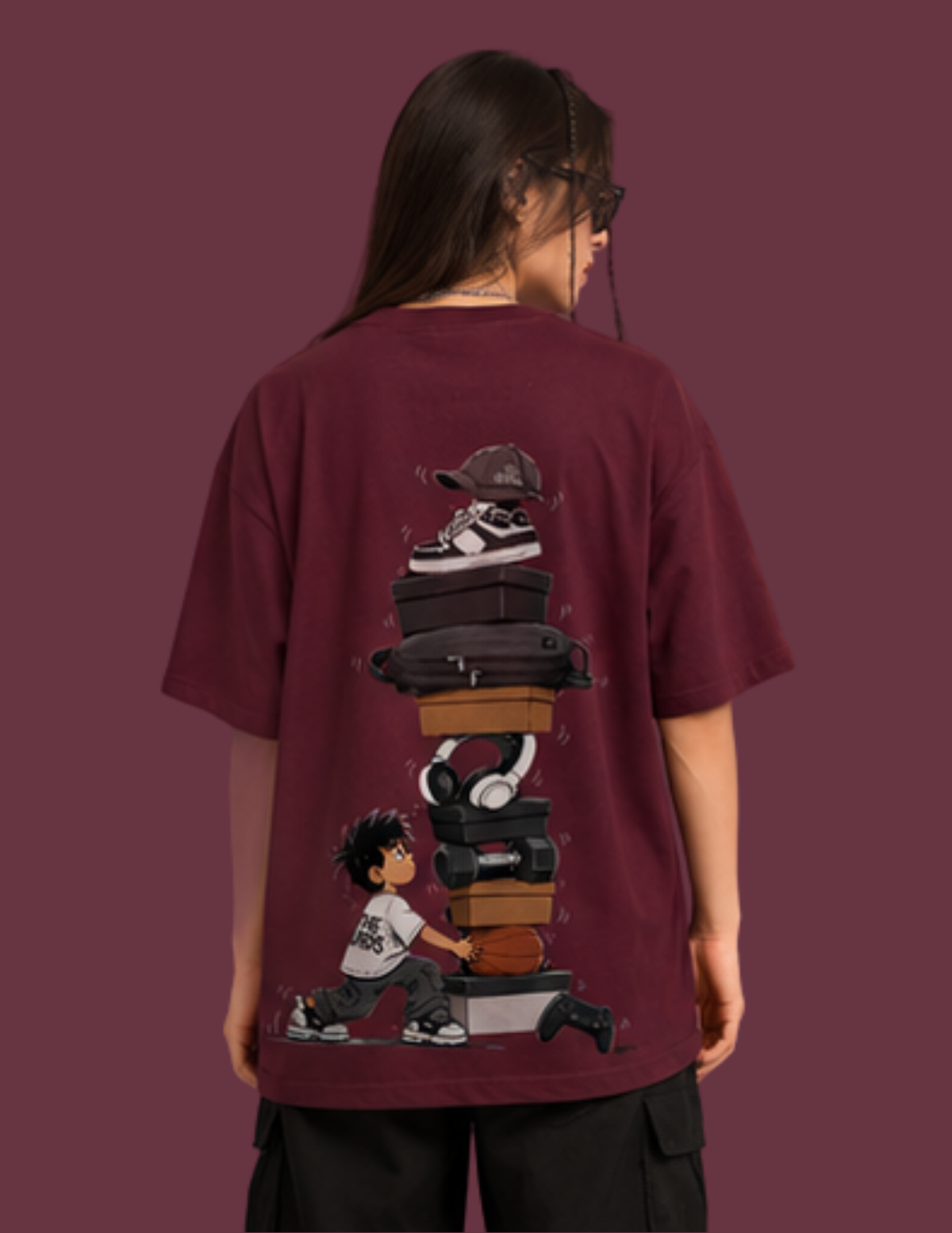 A person shown from the back wearing an oversized maroon t-shirt with a large graphic print on the back. The graphic features a stack of sneakers, headphones, and cases with a cartoon boy pushing a cart.