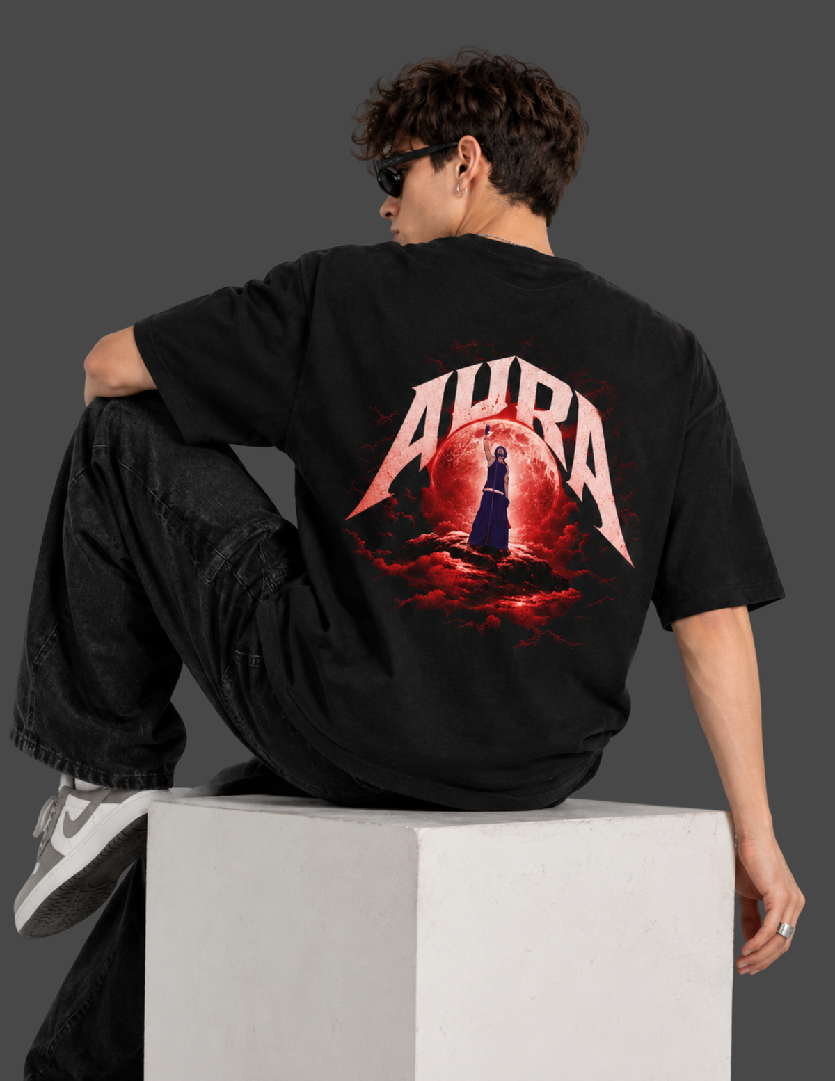 A man wearing a black t-shirt with a large red and white 'AURA' graphic on the back, sitting on a white block against a dark gray background.
