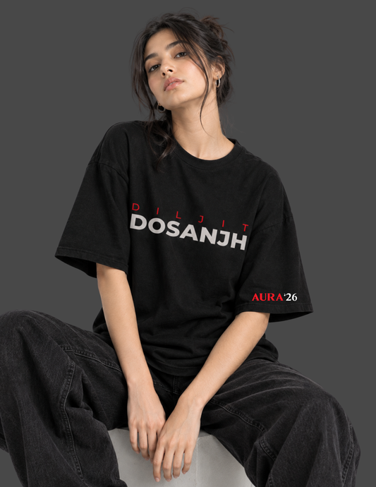 A woman wearing a black oversized t-shirt with 'DOSANJH' in white and red text across the chest, paired with dark gray baggy jeans, sitting against a dark gray background.