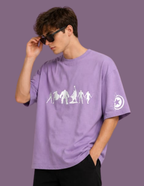 A man wearing a lavender purple oversized t-shirt with a white graphic of game characters on the chest and a moon logo on the sleeve, posed against a solid purple background.