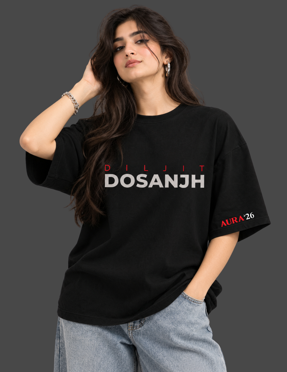 A woman modeling a black oversized graphic T-shirt with 'DILJIT DOSANJH' printed across the chest in white and red, paired with light blue jeans against a dark gray studio background.