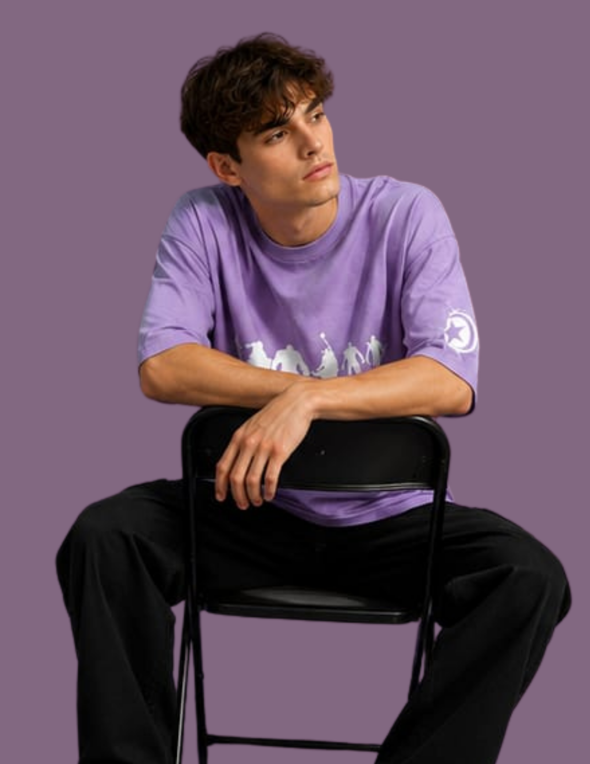 A man sitting on a black folding chair wearing a lavender-purple short-sleeve t-shirt with a white graphic print on the front and a white circular logo on the left sleeve, set against a solid purple background.