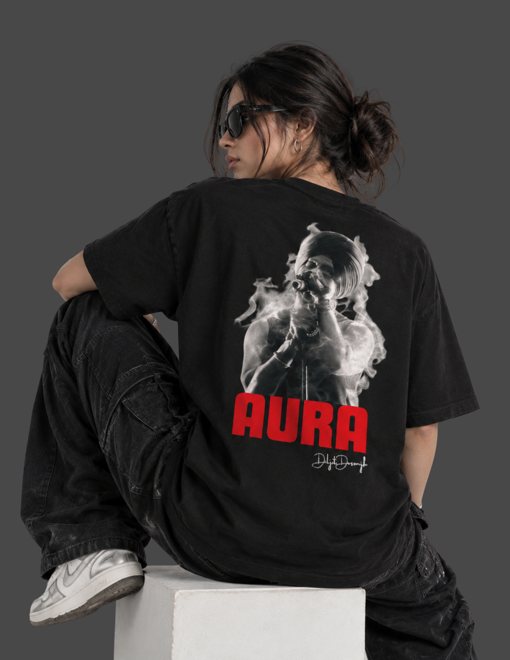 A model wearing a black graphic t-shirt with a large back print of a smoking figure and the text 'AURA' in red, sitting on a white block against a dark gray background.