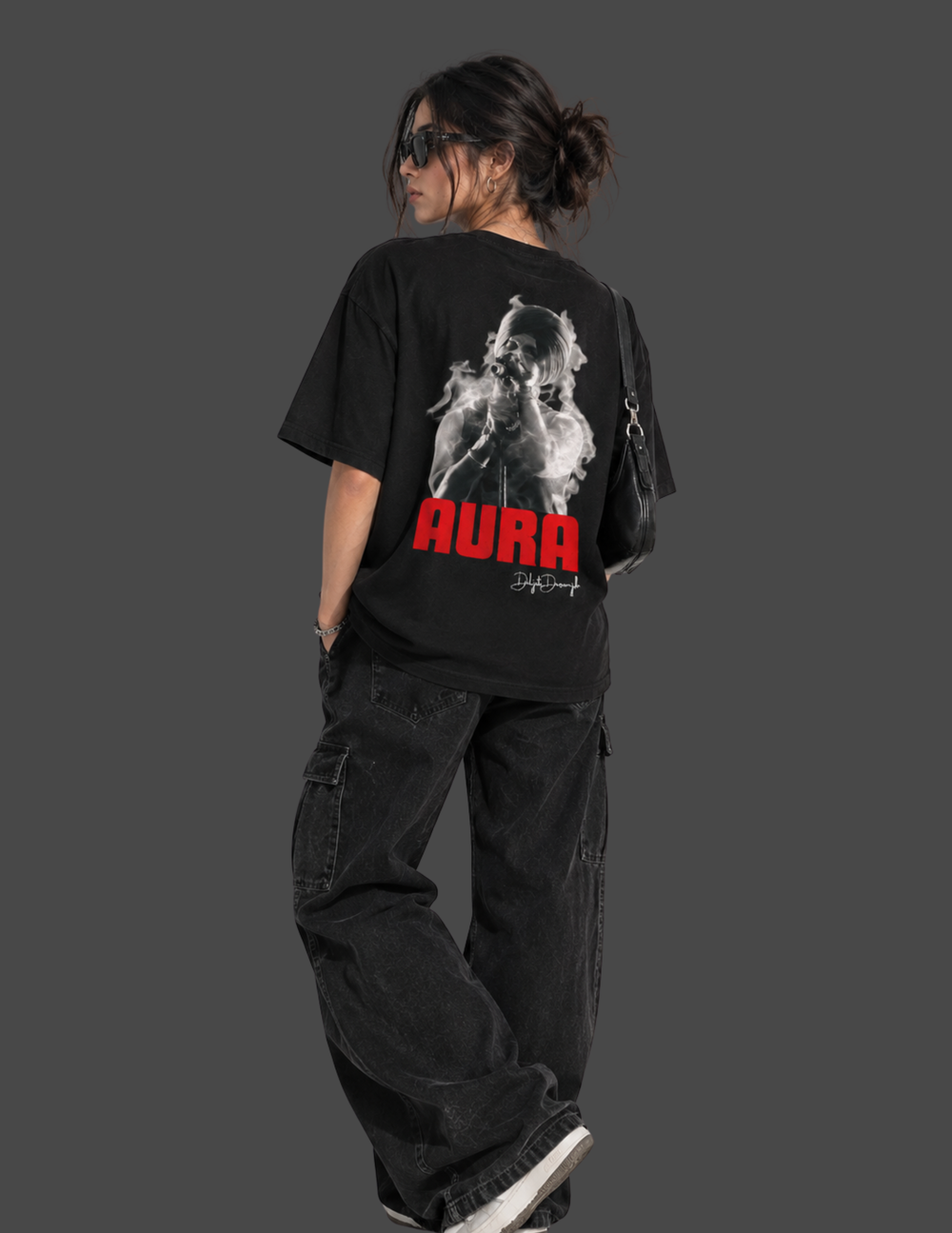 A person shown from the back wearing a black oversized graphic T-shirt with a large white smoky figure print and bold red 'AURA' text, paired with loose black cargo pants against a dark gray studio background.