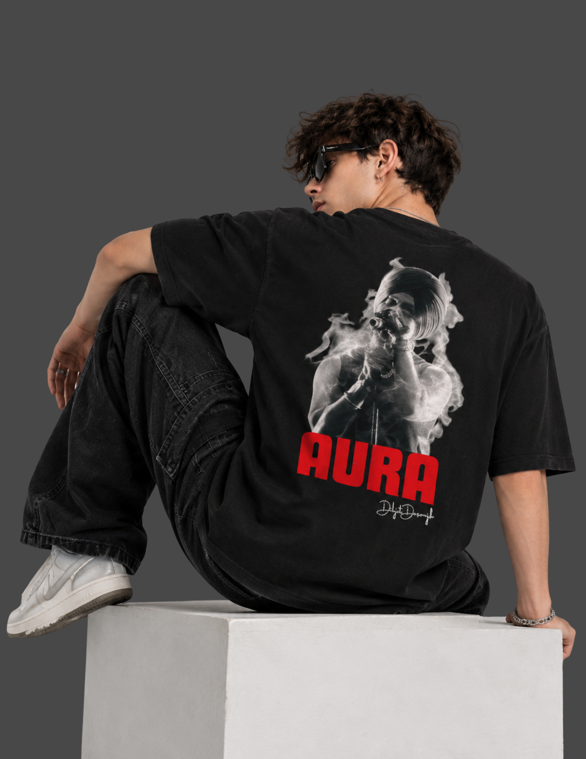 A model wearing a black graphic t-shirt with a large back print of a smoking figure and the word 'AURA' in red, sitting on a white block against a dark gray background.