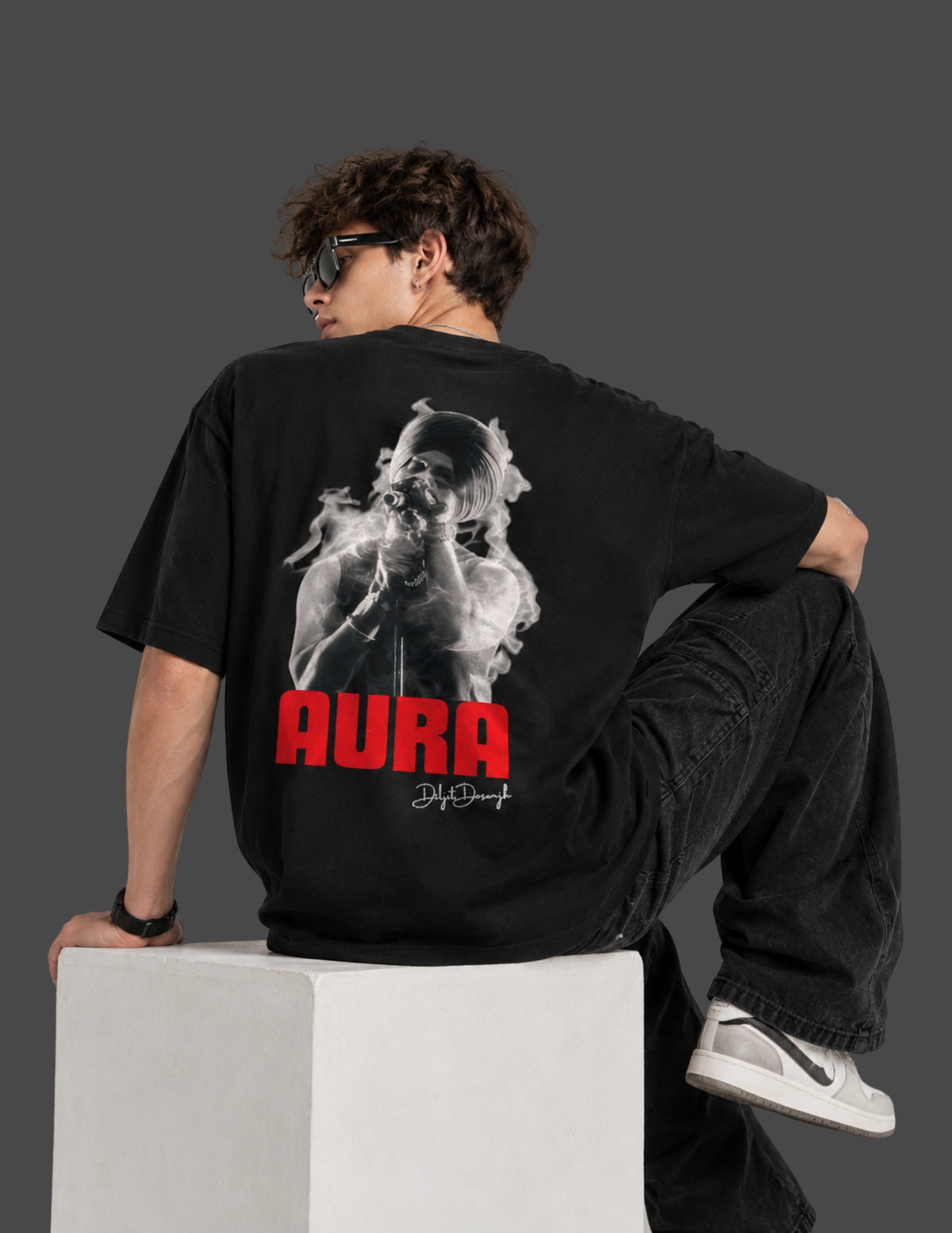 A model wearing a black graphic t-shirt with a large back print of a woman smoking and the word 'AURA' in red, sitting on a white block against a dark gray background.