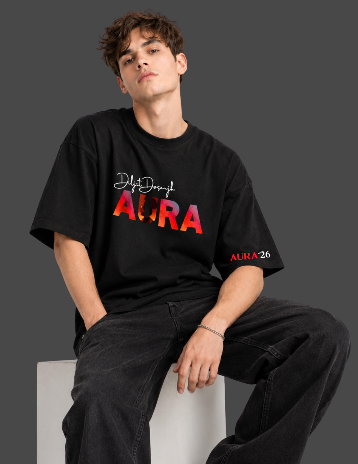 A man wearing a black graphic t-shirt with 'Diyt Dujng AURA' printed on the front and 'AURA26' on the sleeve, paired with black jeans, sitting on a white block against a dark gray background.