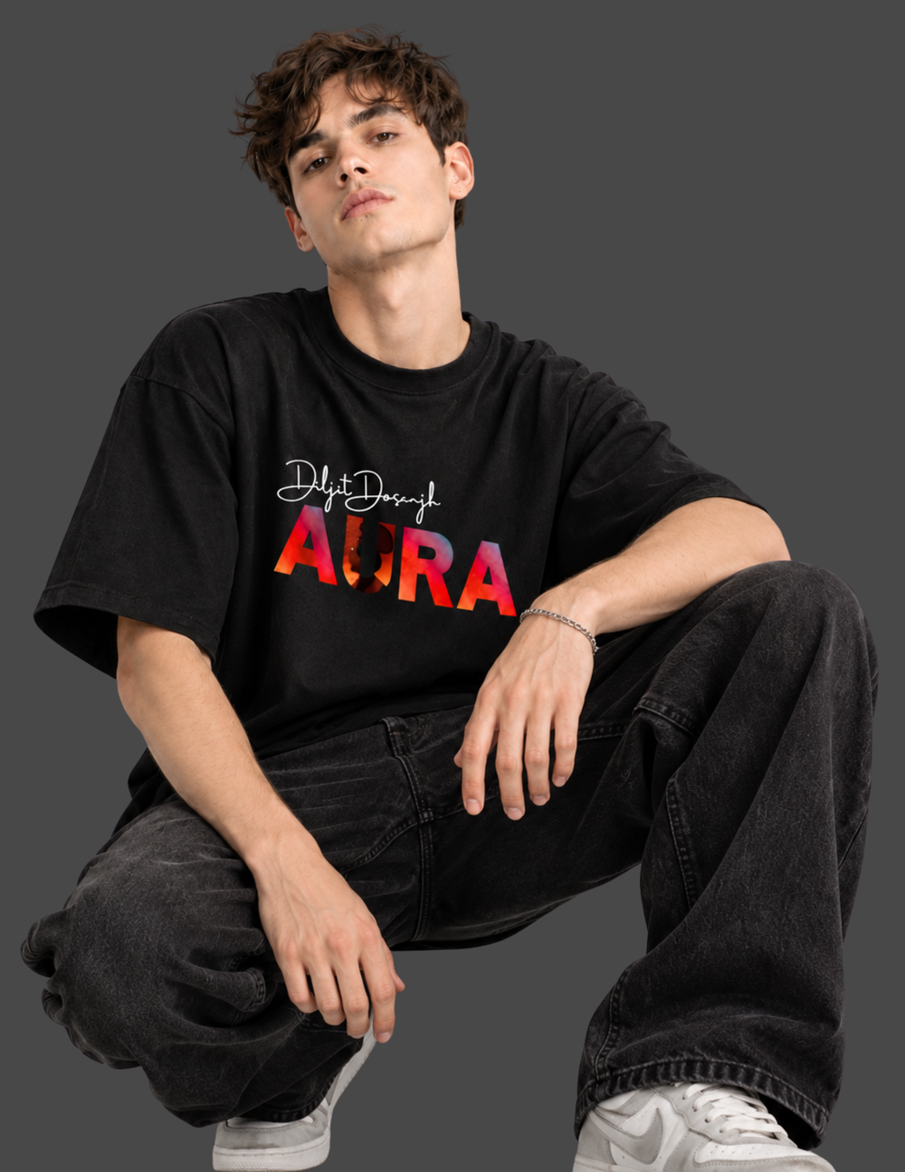 A man wearing a black graphic t-shirt with 'Delhi Delhi AURA' printed on the front, paired with black jeans and white sneakers, posing against a dark gray studio background.
