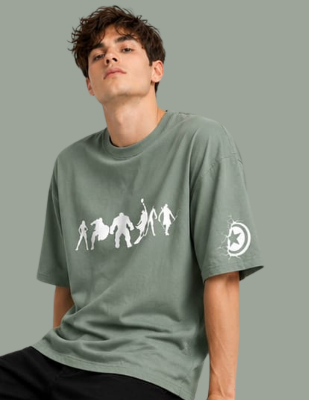 A man wearing an oversized sage green t-shirt with a white graphic of figures on the front and a circular logo on the sleeve, set against a solid sage green background.