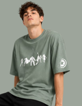 A man wearing an oversized sage green t-shirt with a white graphic of figures on the front and a circular logo on the sleeve, set against a solid sage green background.