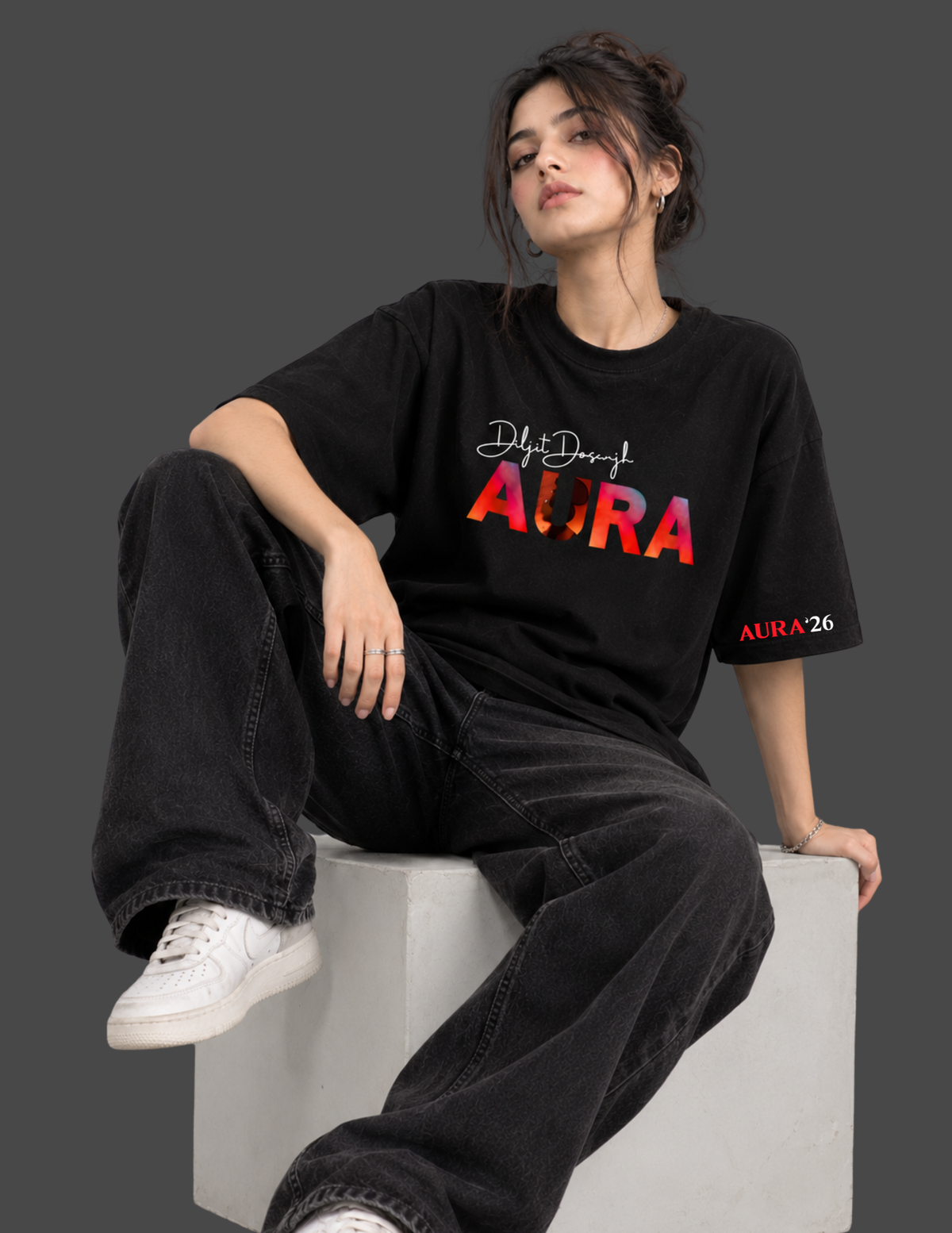 A woman sits on a white block wearing a black graphic t-shirt with 'Aura' written in red and orange gradient letters, paired with black jeans and white sneakers, against a dark gray background.
