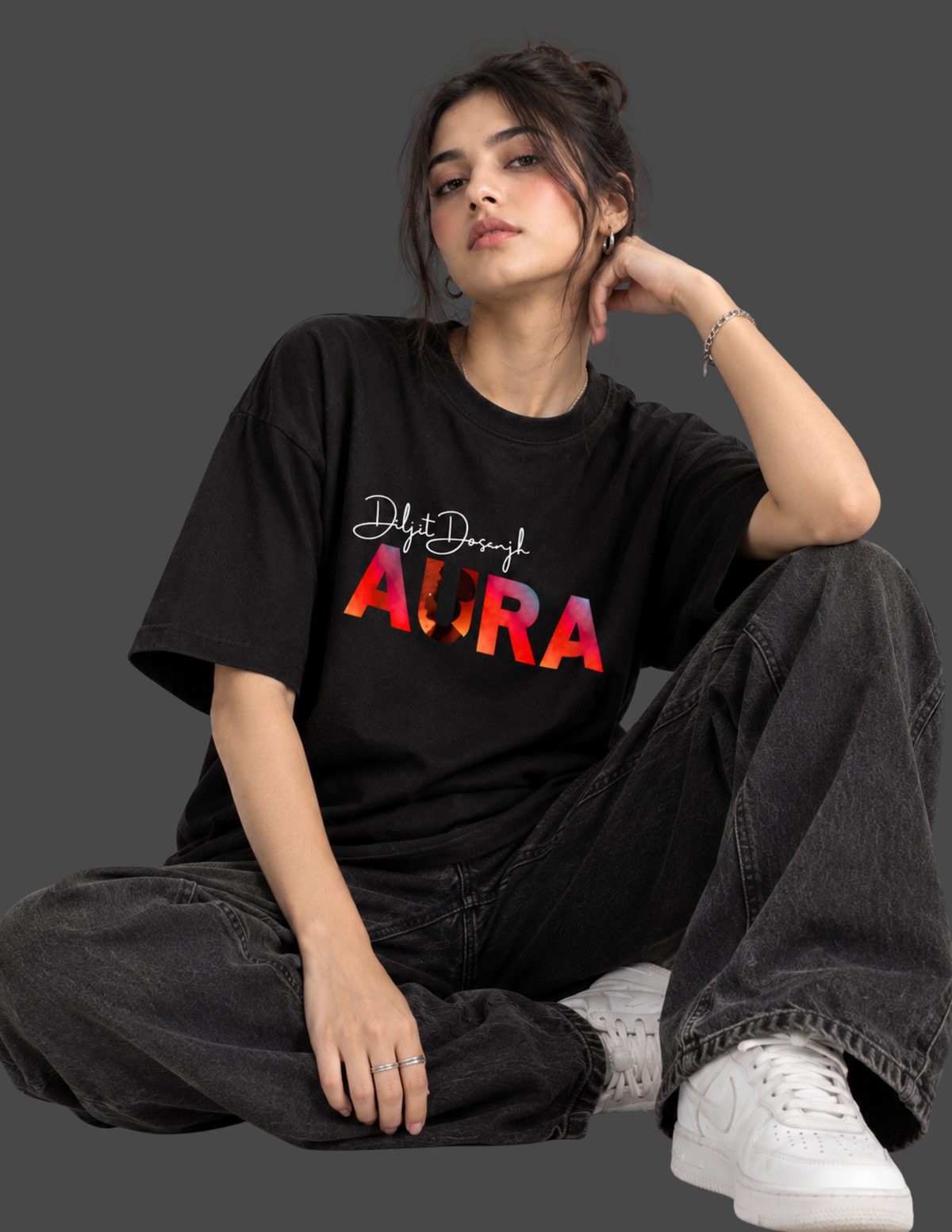 A woman sits cross-legged against a dark gray background wearing a black graphic t-shirt with 'Diljit Dosanjh AORA' printed in white and red gradient text, paired with faded black jeans and white sneakers.