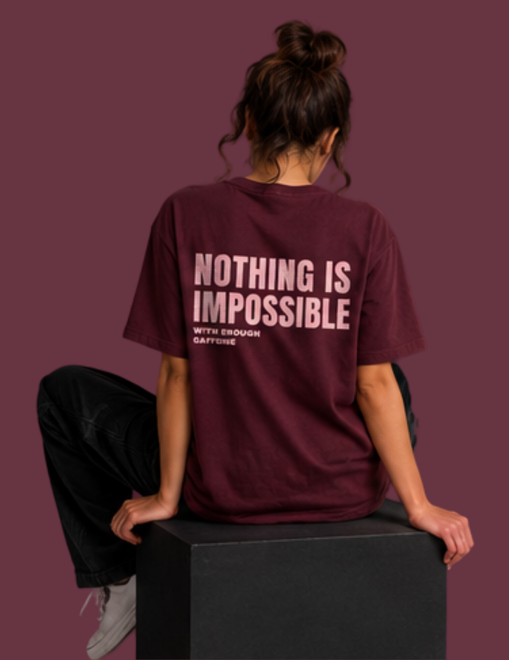 A woman sits on a black block from the back, wearing a maroon t-shirt with the text 'NOTHING IS IMPOSSIBLE WITH ENOUGH CAFECHAINING' printed in light pink on the back. The background is a solid maroon color.