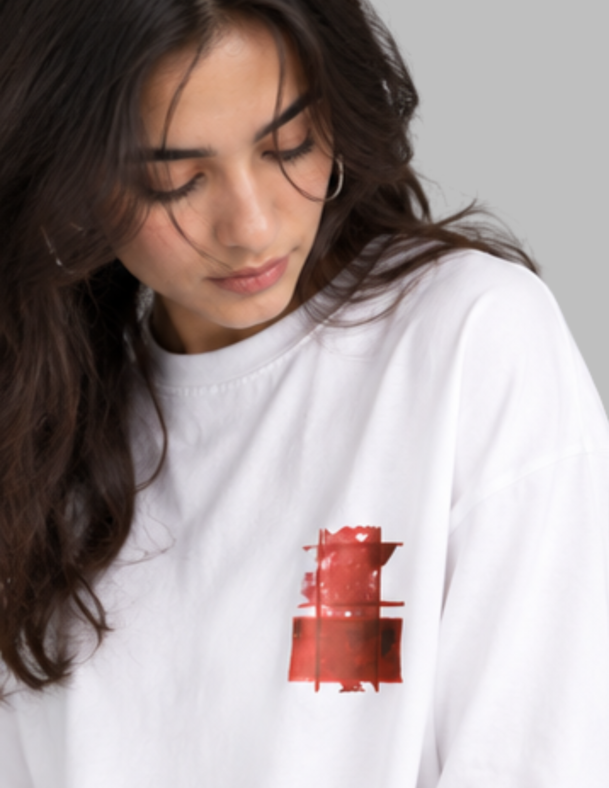 A close-up of a woman wearing a white crewneck sweatshirt with a red graphic print on the left chest, set against a solid gray background.