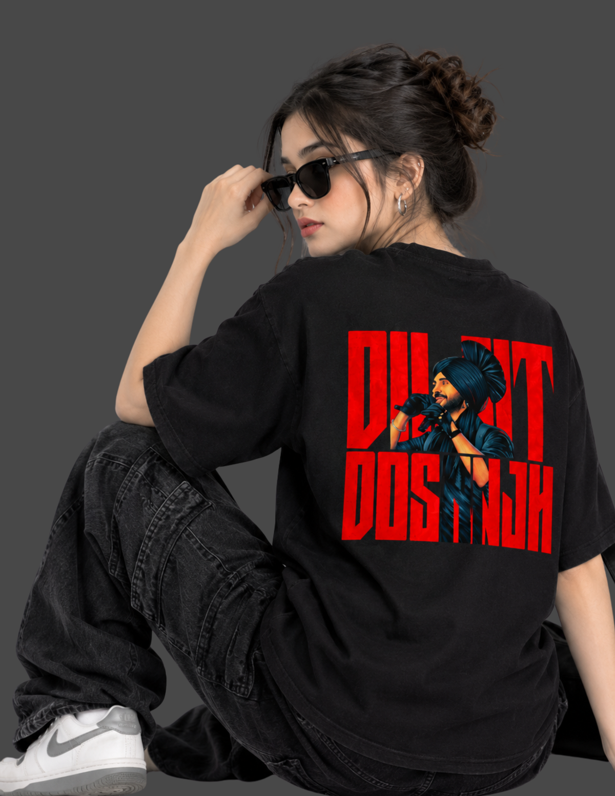 A woman wearing a black oversized t-shirt with a bold red and blue graphic print on the back, paired with dark gray cargo pants and white sneakers, posed against a solid dark gray studio background.