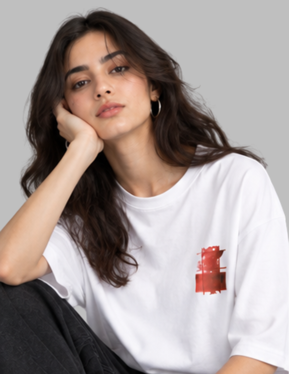 A woman with long dark hair rests her head on her hand, wearing a white short-sleeve t-shirt with a small red graphic on the left chest, set against a solid gray background.