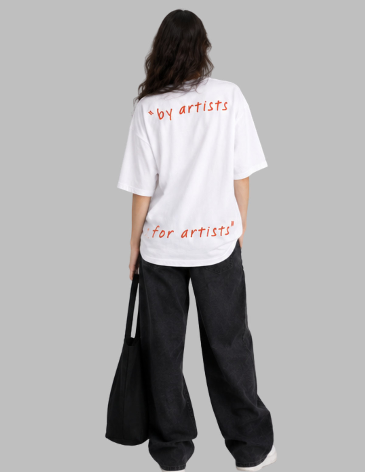A model shown from the back wearing a white oversized t-shirt with red text reading '+by artists' on the upper back and 'for artists' on the lower back, paired with black wide-leg pants and a black tote bag, against a solid gray background.