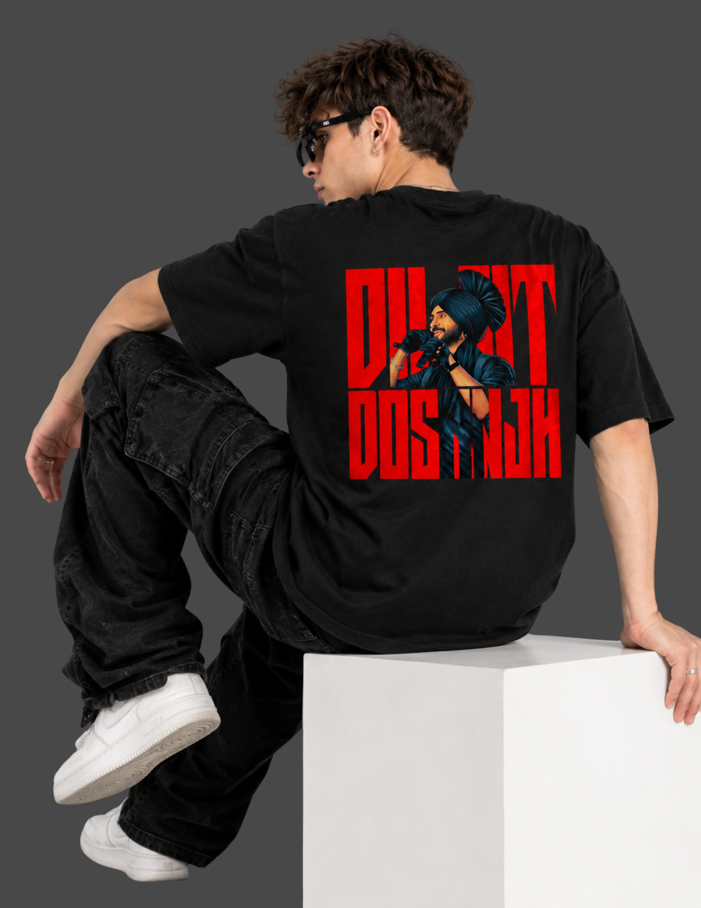 A man wearing a black t-shirt with a bold red graphic print of 'DILYT DOS HAH' and a character illustration, sitting on a white block against a dark gray background.