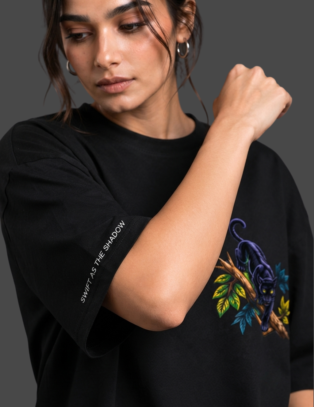 A woman wearing a black oversized t-shirt with a colorful graphic of a panther on a branch and white text on the sleeve, set against a dark gray background.
