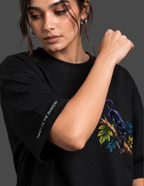 A woman wearing a black oversized t-shirt with a colorful graphic of a panther on a branch and white text on the sleeve, set against a dark gray background.