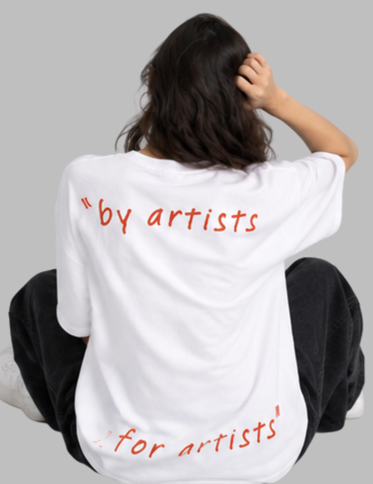 A person sitting with their back to the camera wearing a white oversized T-shirt with red handwritten-style text reading "by artists for artists" on a plain gray background.