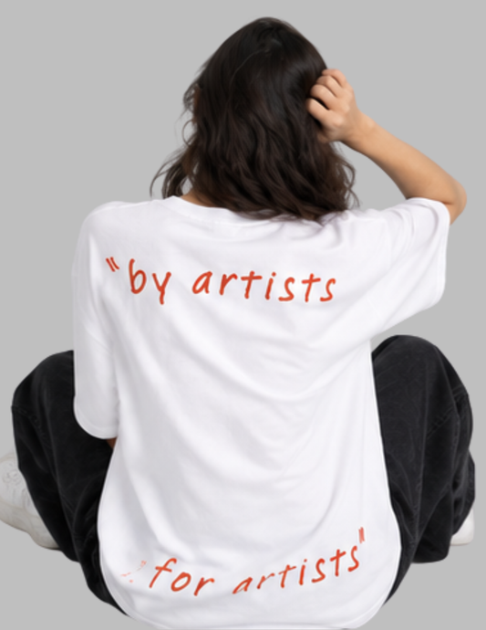 A person sitting with their back to the camera wearing a white oversized T-shirt with red handwritten-style text reading "by artists for artists" on a plain gray background.