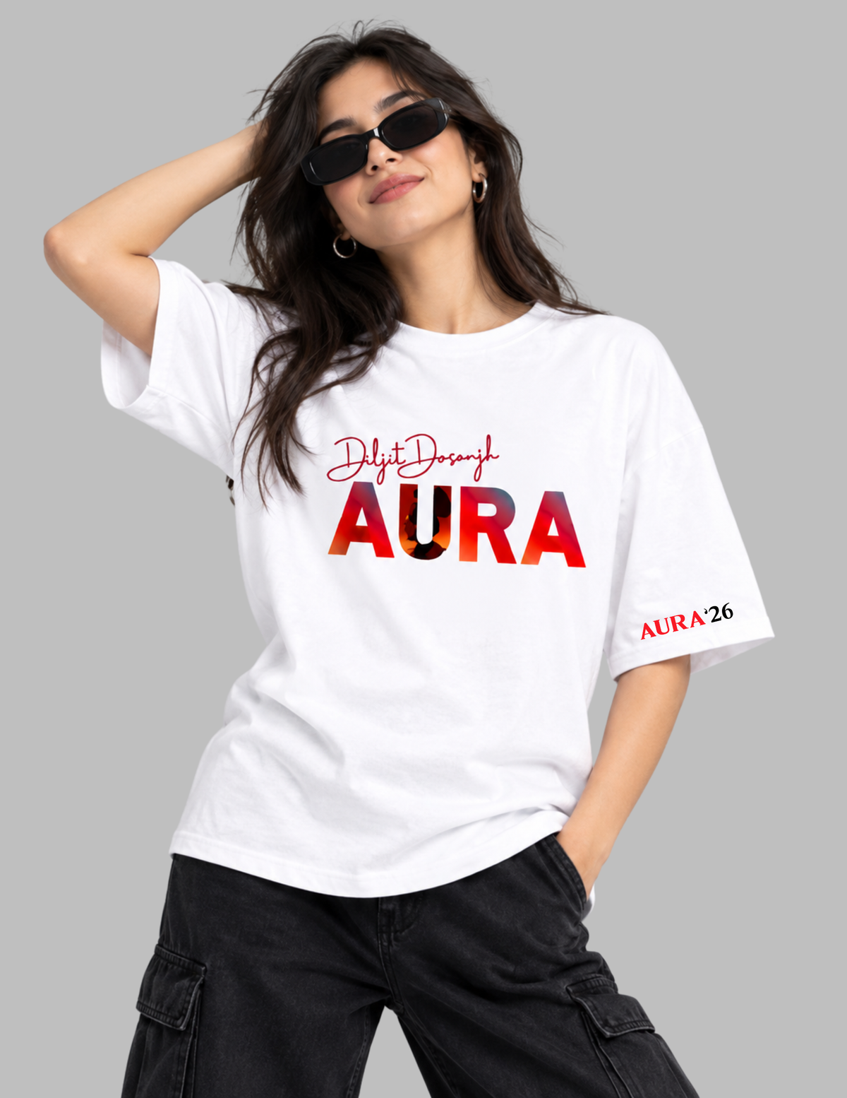 A woman wearing a white graphic t-shirt with 'Delhi Through AURA 26' printed on the front, paired with black cargo pants and sunglasses, set against a solid gray background.