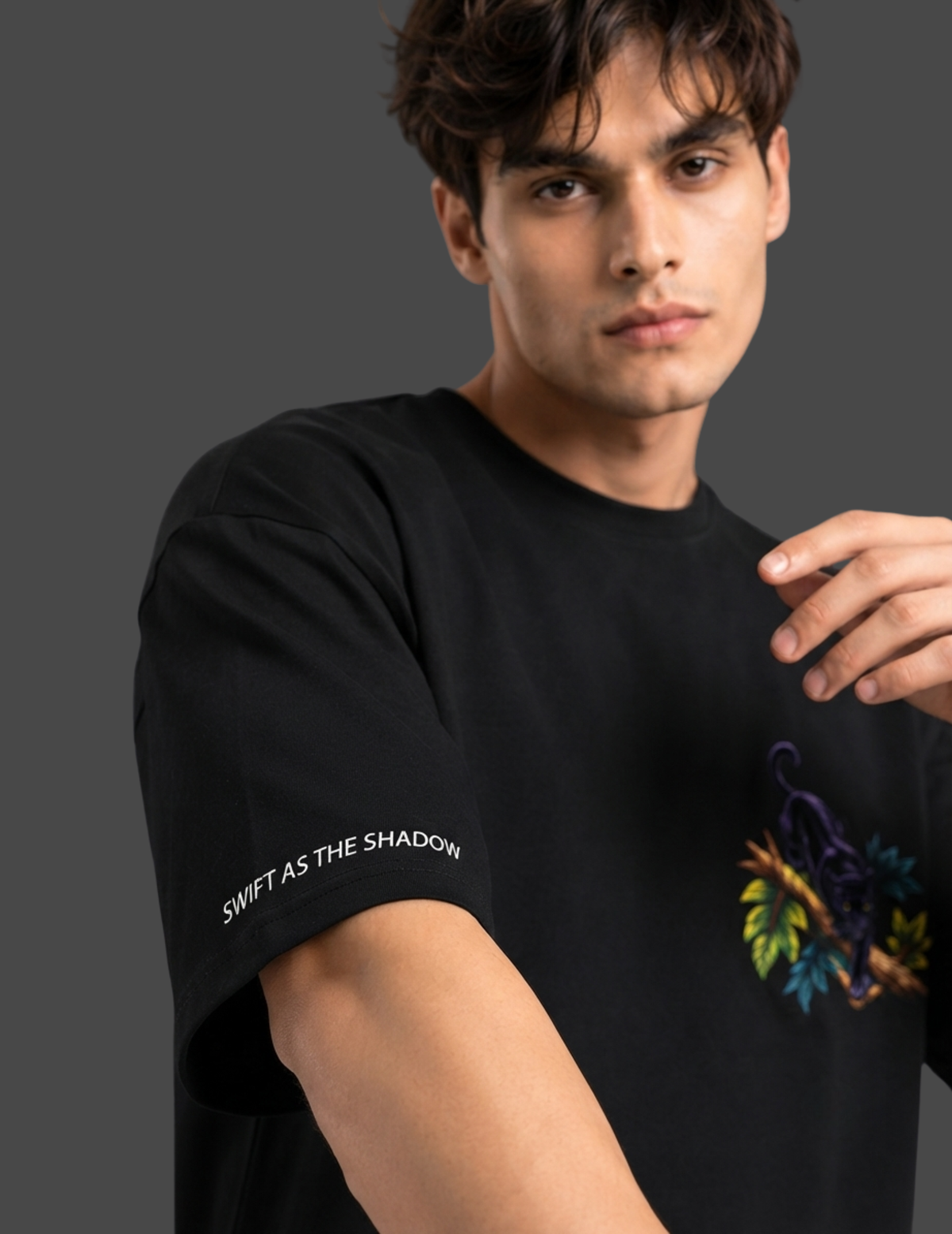 A man wearing a black short-sleeve t-shirt with white text on the sleeve reading 'SWIFT AS THE SHADOW' and a colorful graphic on the chest, photographed against a dark gray studio background.