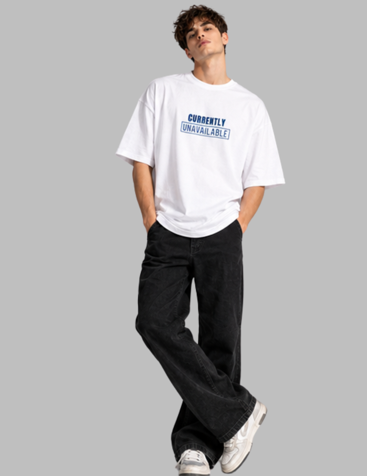 A man modeling a white oversized t-shirt with a blue graphic text print that reads 'CURRENTLY UNAVAILABLE', paired with black jeans and white sneakers, set against a solid gray background.