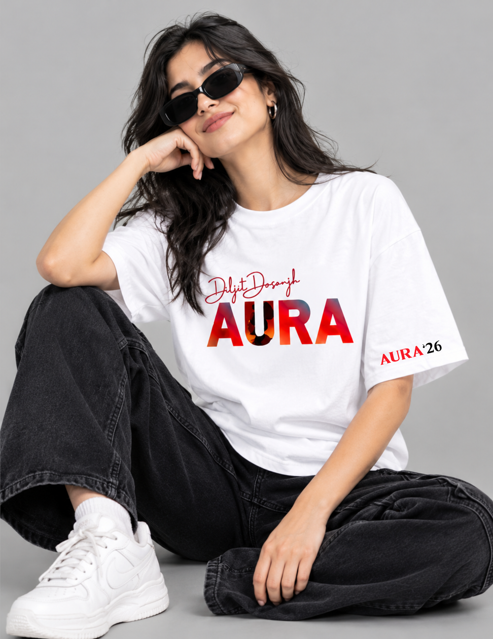A woman wearing a white graphic t-shirt with 'DilysDesign AURA' printed on the front, paired with black jeans and white sneakers, sitting against a solid gray background.