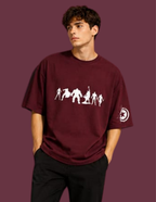 A man wearing an oversized maroon t-shirt with a white graphic of human evolution silhouettes on the front and a circular logo on the sleeve, set against a solid maroon background.