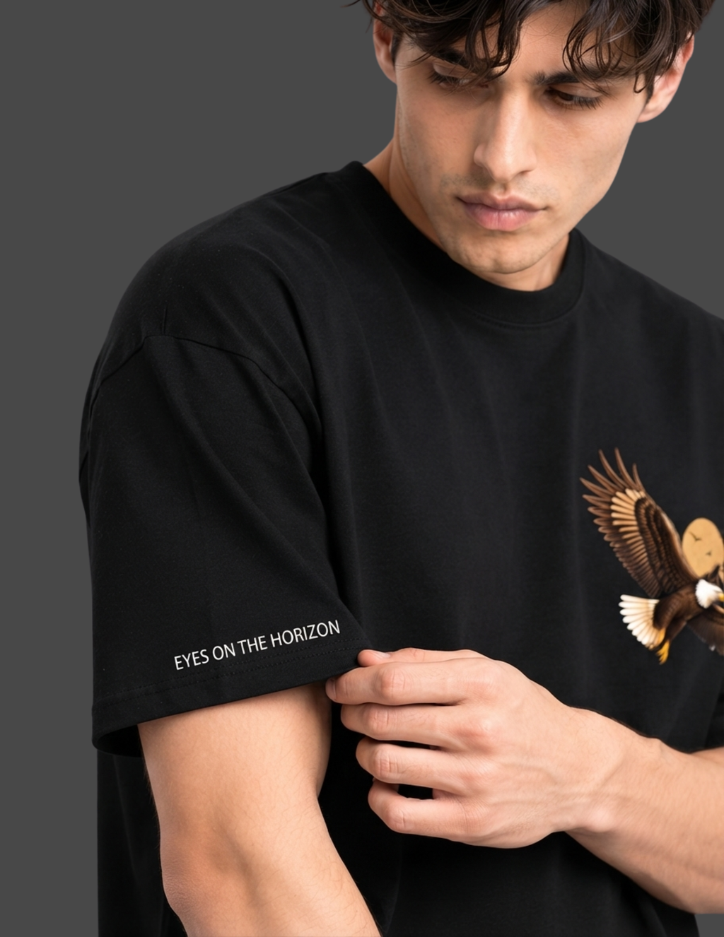 A man wearing a black short-sleeve t-shirt with a graphic of an eagle on the right chest and the text 'EYES ON THE HORIZON' printed in white on the left sleeve, set against a dark gray background.
