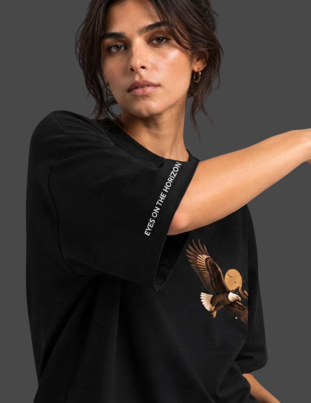 A woman wearing a black oversized t-shirt with a graphic of an eagle holding a sun on the front and white text on the sleeve, set against a dark gray background.
