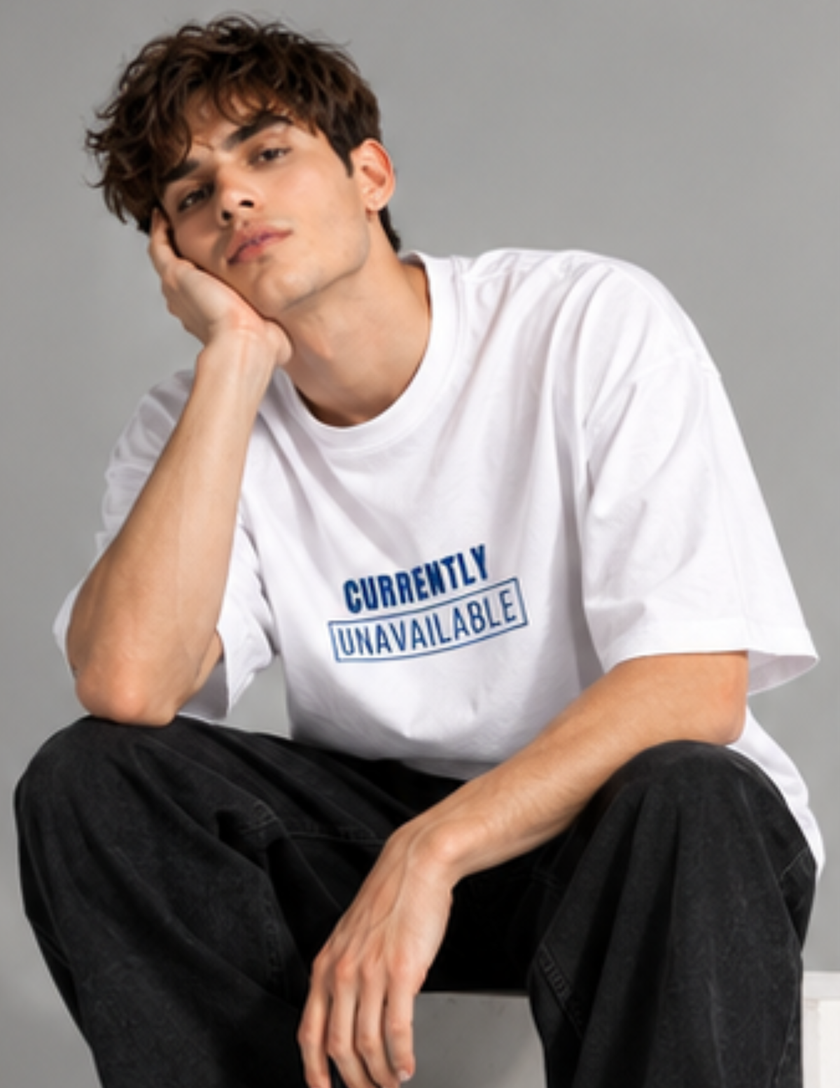 A young man with wavy brown hair sits in a studio setting, wearing a white graphic t-shirt with the text 'CURRENTLY UNAVAILABLE' printed in blue on the chest, paired with black jeans.