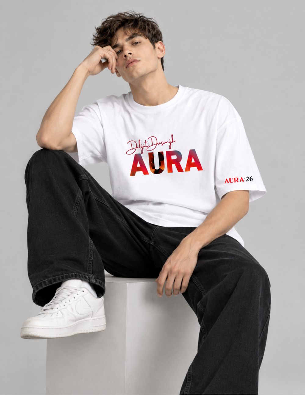 A man wearing a white t-shirt with a red and black graphic print that reads 'DiyfDiyf AURA' and 'AURA 26' on the sleeve, paired with black jeans and white sneakers, sitting on a white block against a gray background.