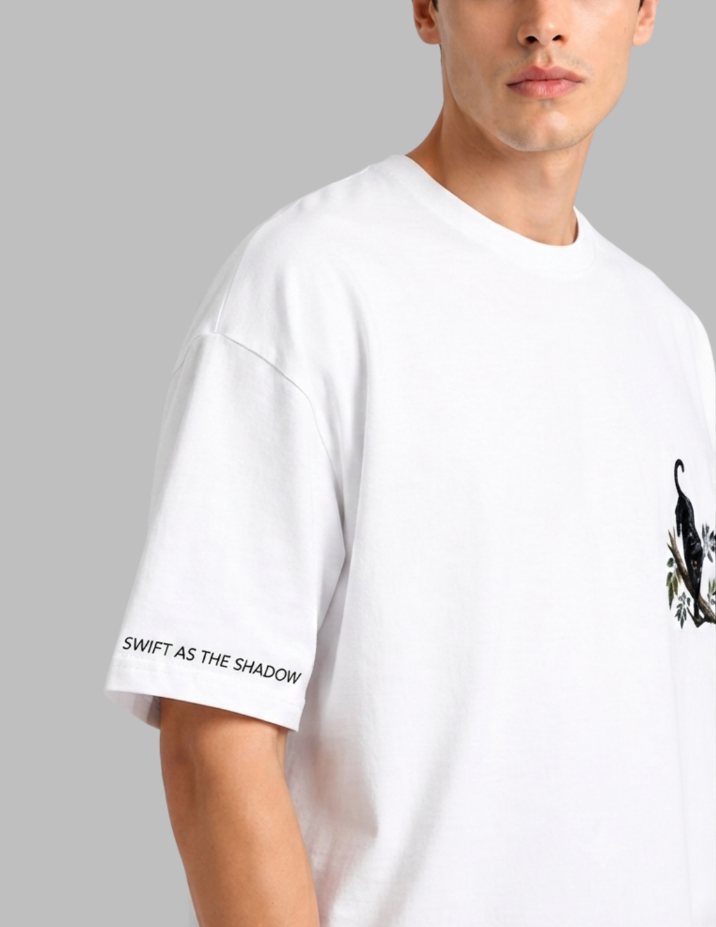 A close-up of a model wearing a white oversized crewneck t-shirt. The right sleeve features the black text 'SWIFT AS THE SHADOW', and the right chest area has a black graphic of a bird perched on a leafy branch.