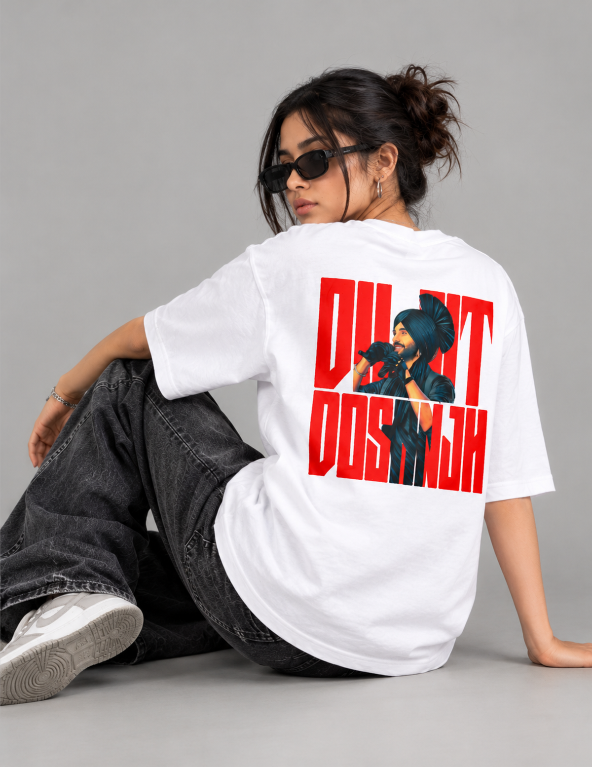 A model wearing a white oversized graphic t-shirt with a bold red and black print on the back, paired with dark gray jeans and white sneakers, sitting against a solid gray studio background.