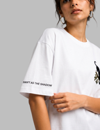 A close-up of a model wearing an oversized white t-shirt. The shirt features the text 'SWIFT AS THE SHADOW' printed in black on the left sleeve and a black bird graphic on the right chest.