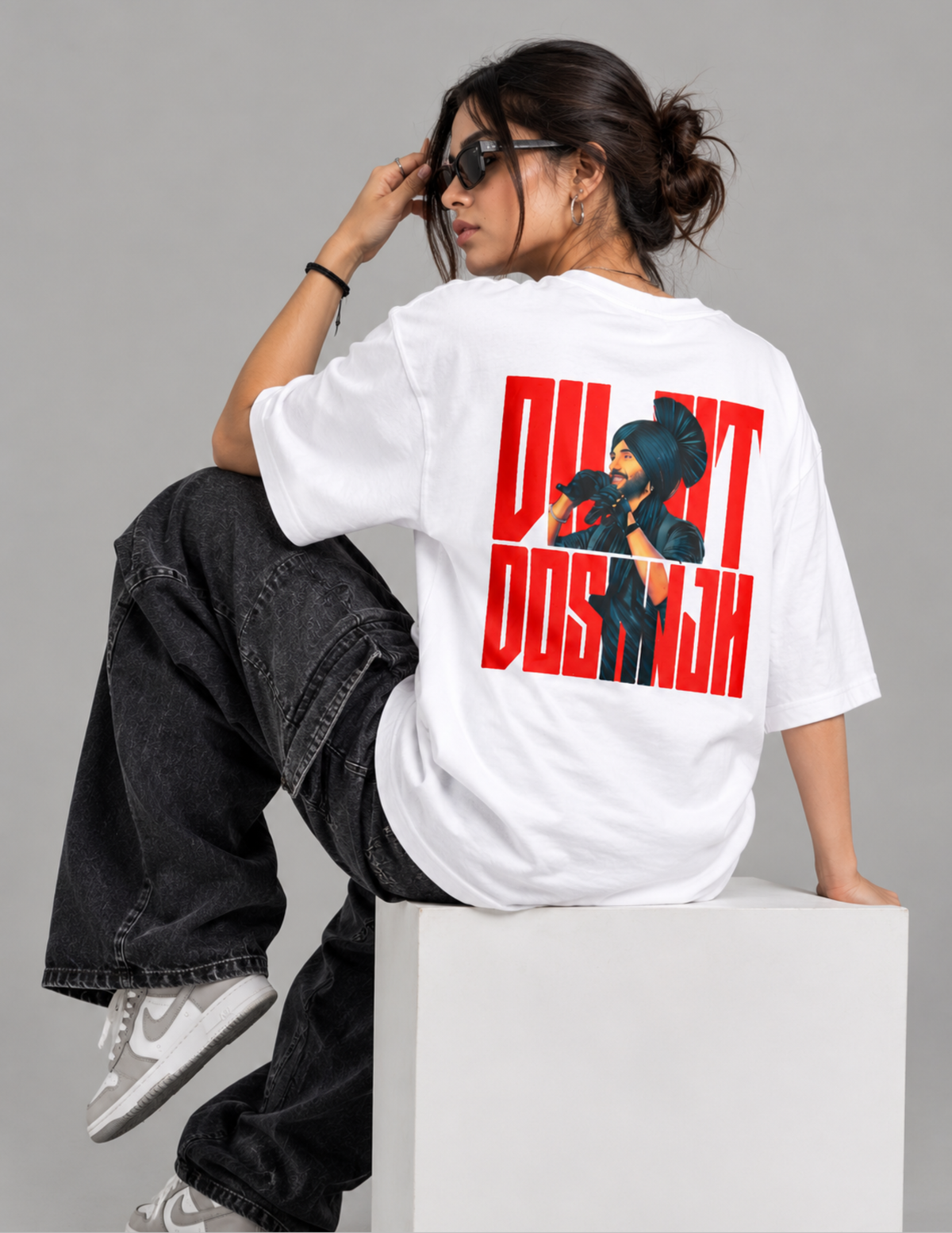 A woman wearing a white oversized t-shirt with a bold red and black graphic print on the back, paired with black jeans and white sneakers, sitting on a white block against a gray studio background.