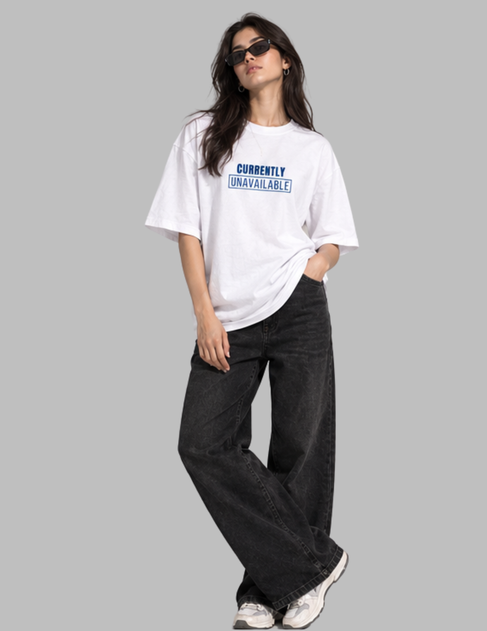 A woman modeling a white oversized graphic t-shirt with blue text that reads 'CURRENTLY UNAVAILABLE!' paired with dark wide-leg jeans and white sneakers, set against a solid gray background.