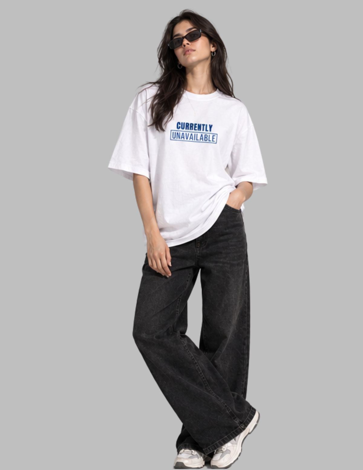 A woman modeling a white oversized graphic t-shirt with blue text that reads 'CURRENTLY UNAVAILABLE!' paired with dark wide-leg jeans and white sneakers, set against a solid gray background.
