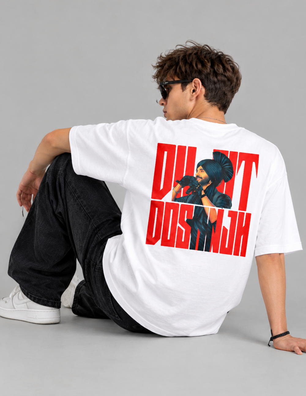 A man wearing a white oversized T-shirt with a large red and black graphic print on the back, paired with black jeans and white sneakers, seated against a light gray studio background.