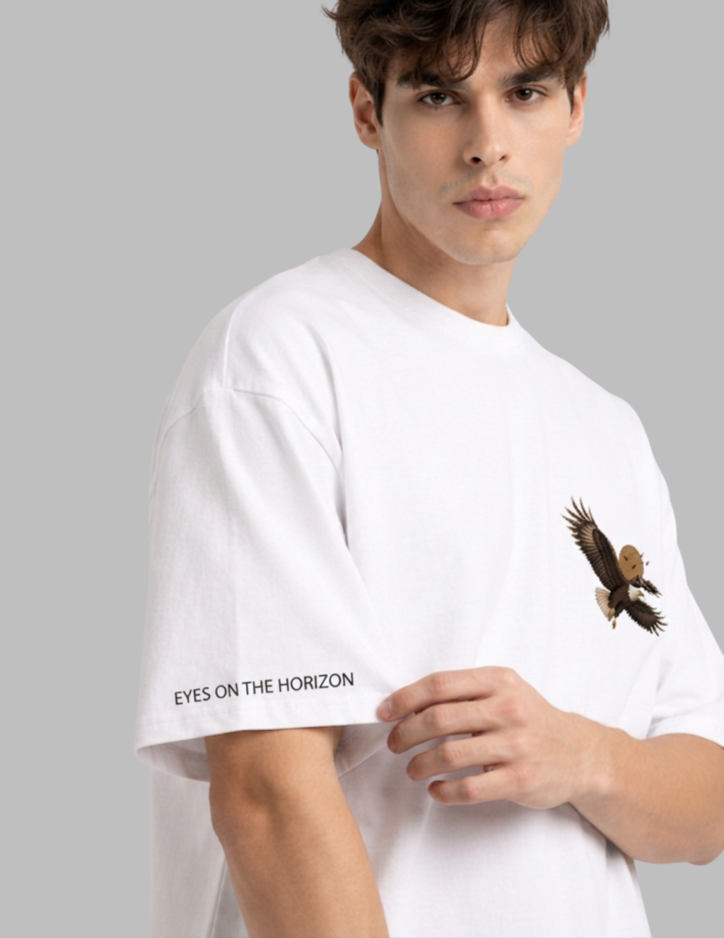 A man wearing a white crewneck t-shirt with a graphic of a flying eagle on the left chest and the text 'EYES ON THE HORIZON' printed on the right sleeve, set against a solid gray background.