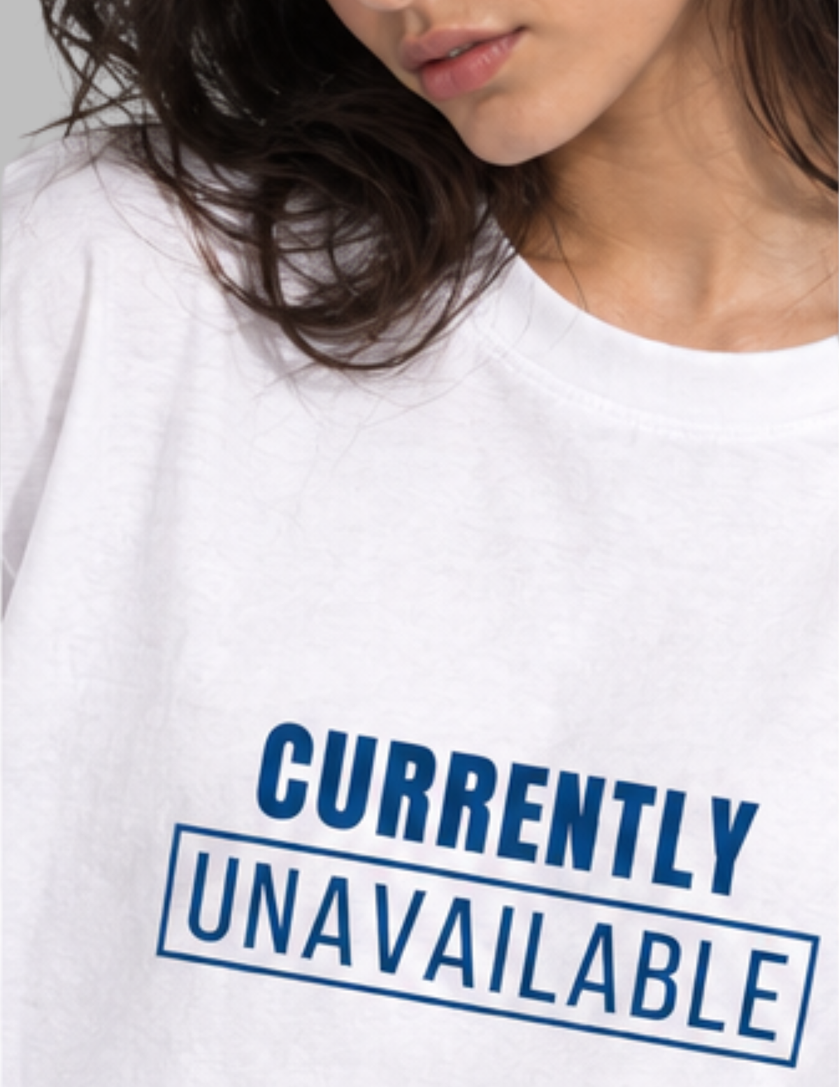 A close-up of a person wearing a white crewneck T-shirt with a blue graphic print that reads 'CURRENTLY UNAVAILABLE' inside a rectangular border.