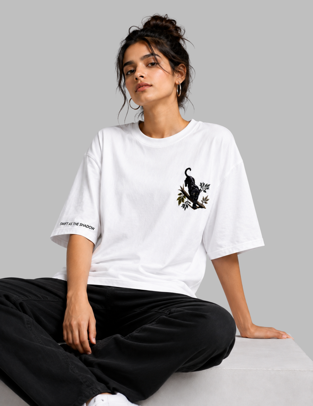 A woman sits on a light gray surface wearing an oversized white T-shirt with a black cat graphic on the left chest and black pants, photographed against a plain light gray studio background.