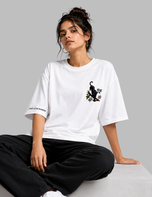 A woman sits on a light gray surface wearing an oversized white T-shirt with a black cat graphic on the left chest and black pants, photographed against a plain light gray studio background.