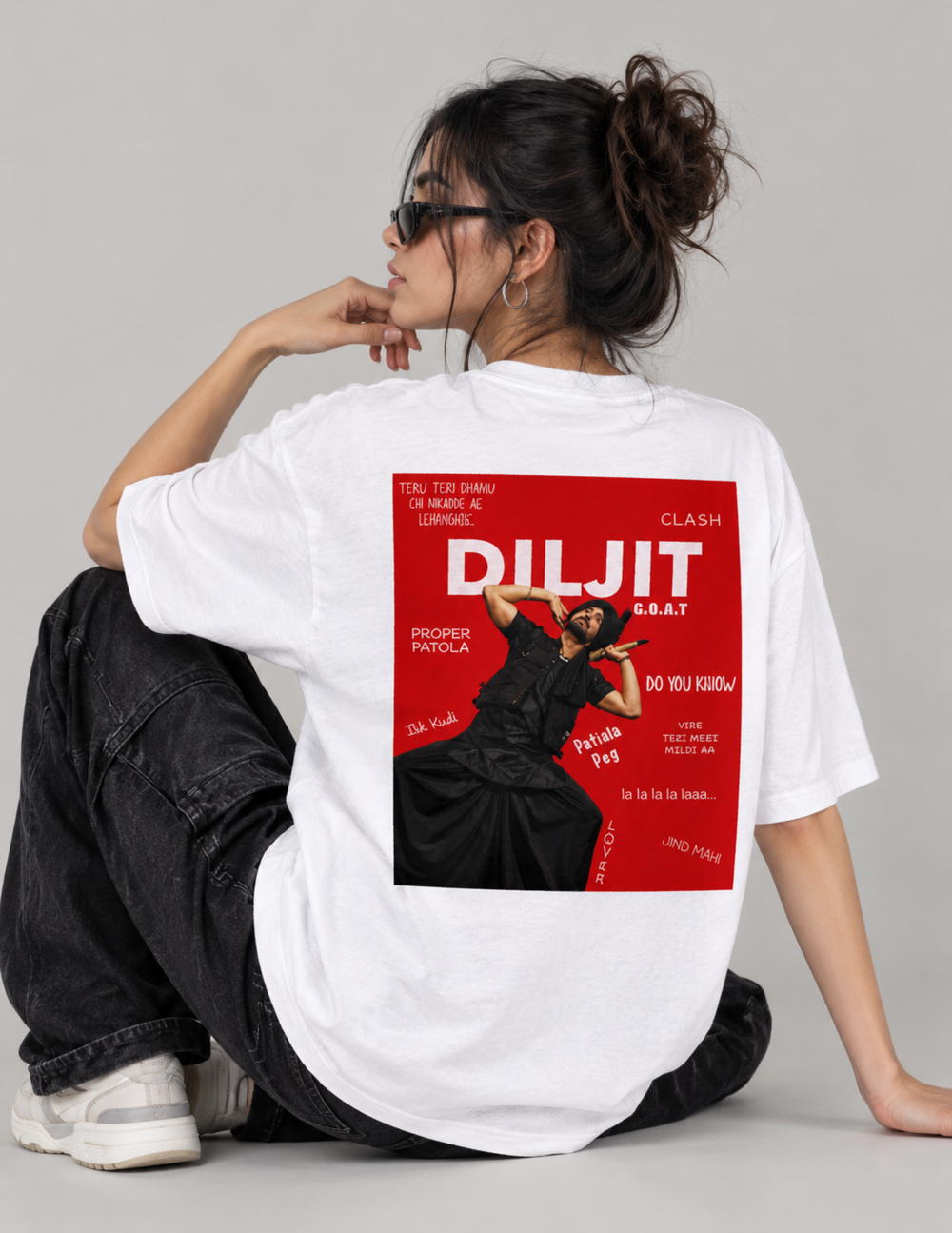 A woman sits on the floor wearing a white oversized T-shirt with a large red and black graphic print on the back, paired with dark jeans and white sneakers against a light gray studio background.